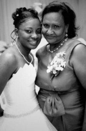 Two women posing for a photo, one in a white dress with a pearl necklace and the other in a light-colored dress with a floral corsage.