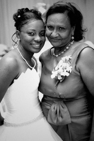 Two women posing for a photo, one in a white dress with a pearl necklace and the other in a light-colored dress with a floral corsage.